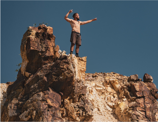 Shirtless man on top of mountain with hands in air, symbolizing persistence.
